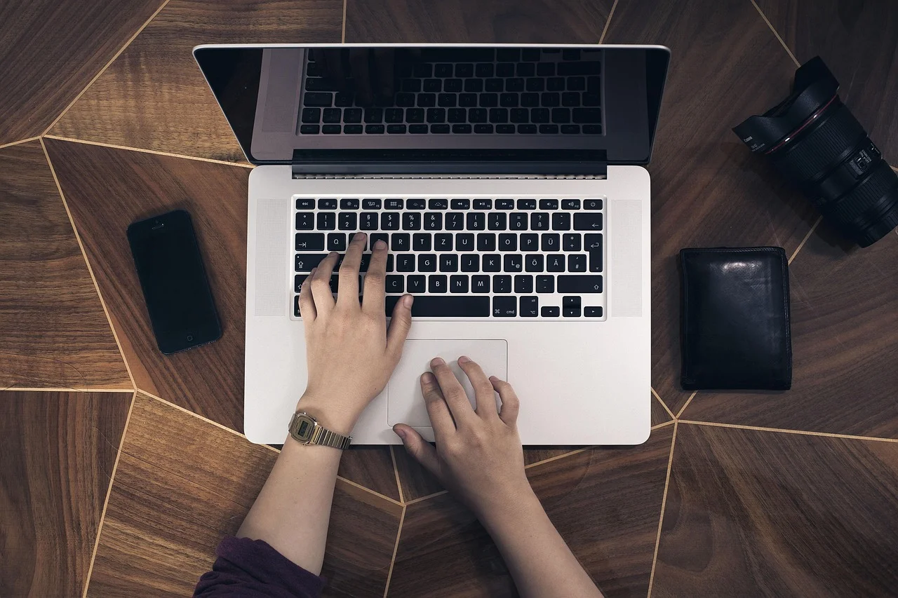 Personne remplissant un formulaire de demande en ligne sur un ordinateur portable, téléphone et portefeuille posés à côté du clavier.