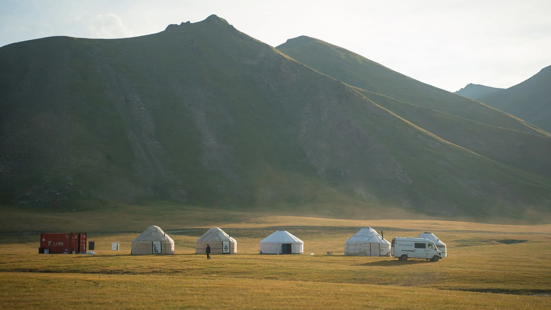 Rangée de yourtes blanches sur un pâturage d'altitude kirghize à l'aube, adossée à une montagne verte, avec un van et un berger marchant devant les tentes