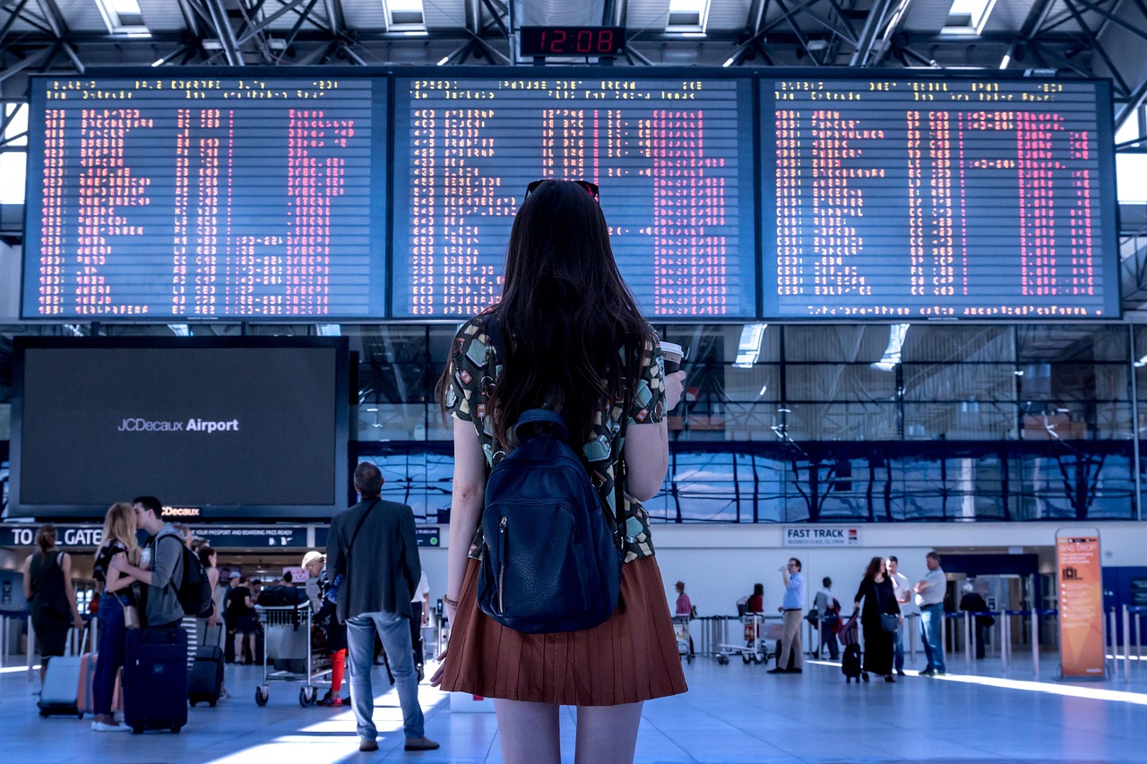 Voyageuse face au tableau des départs d'un aéroport, sous un panneau rappelant de préparer passeport et carte d'embarquement.