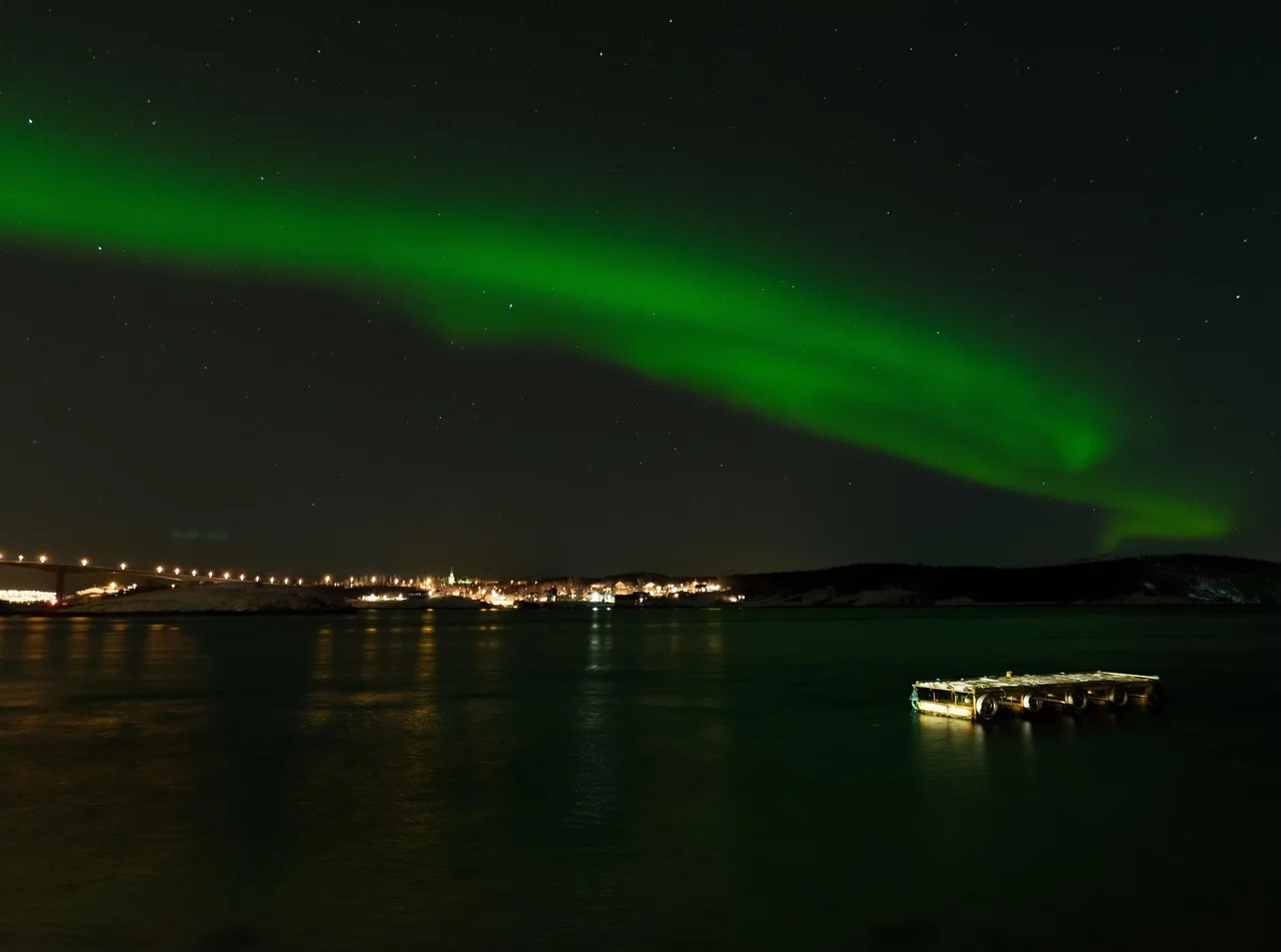 Aurore boréale verte au-dessus des lumières de la ville de Tromsø et de son pont, vues depuis la rive opposée du détroit.