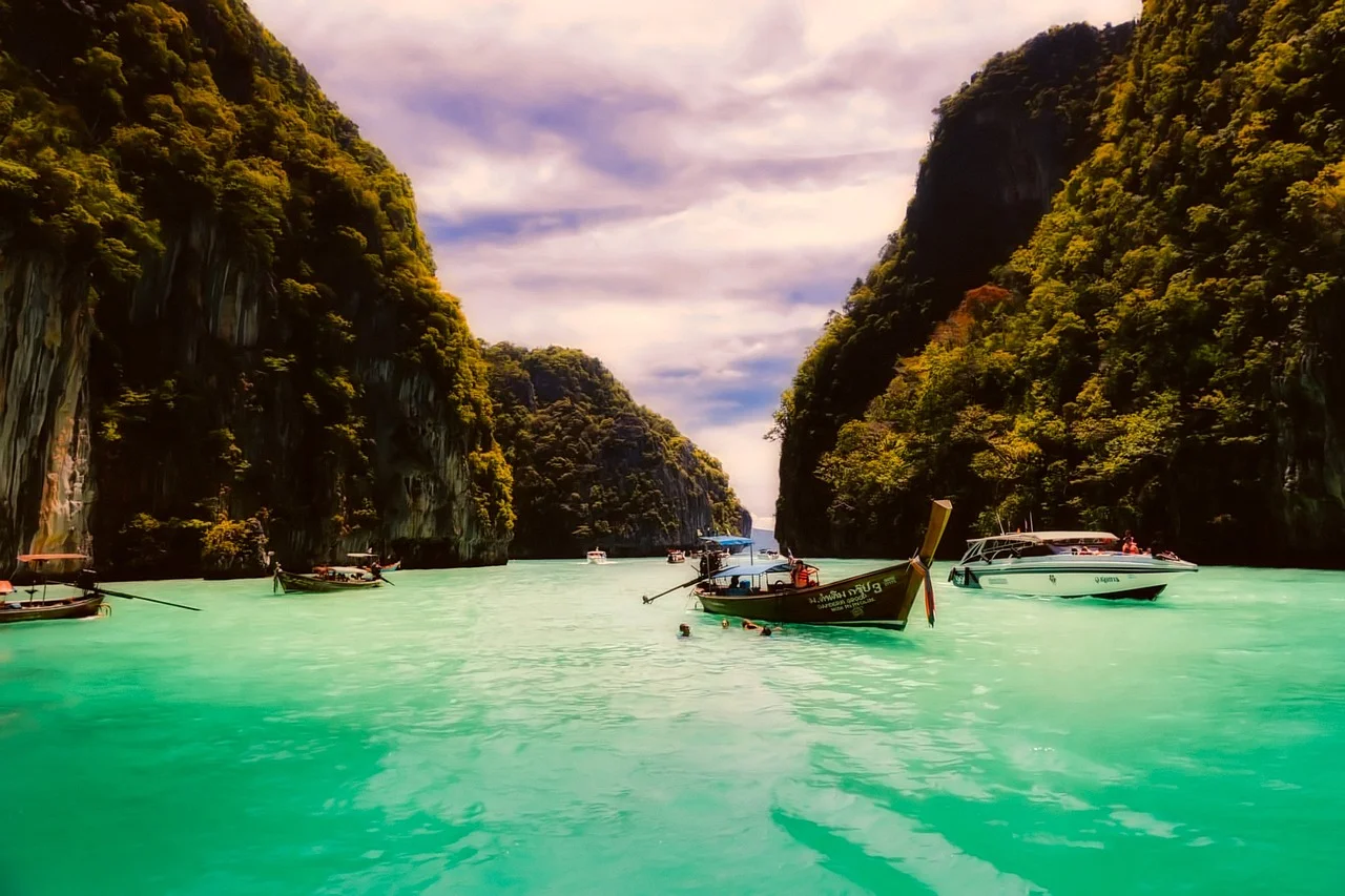 Bateaux longtail thaïlandais amarrés dans une baie d'eau turquoise encadrée de hautes falaises calcaires couvertes de végétation, mer d'Andaman, sud de la Thaïlande.