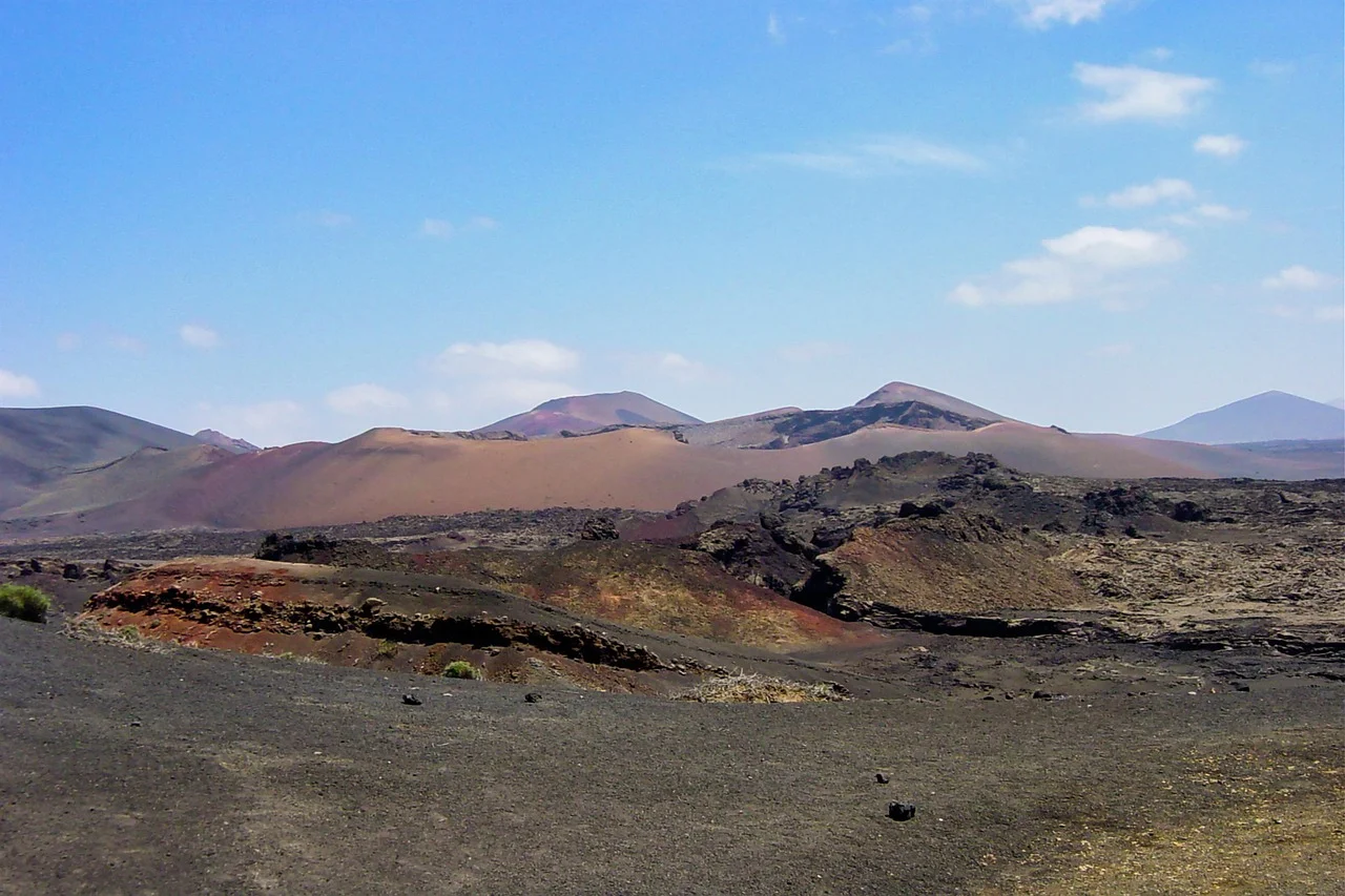 Cônes volcaniques ocre et rouge du parc national de Timanfaya à Lanzarote sous un ciel bleu, champ de lave noire au premier plan