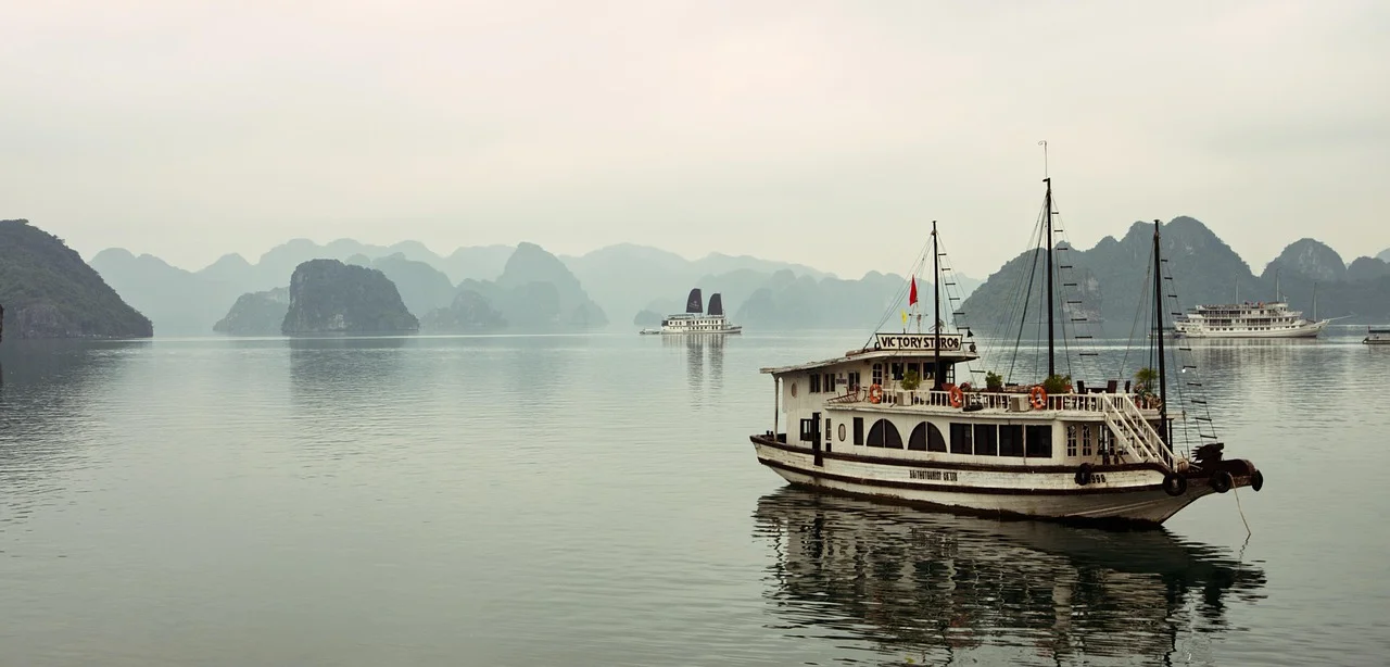 Jonque en bois blanche voguant sur les eaux calmes de la baie d'Ha Long, devant des pitons karstiques estompés par la brume.