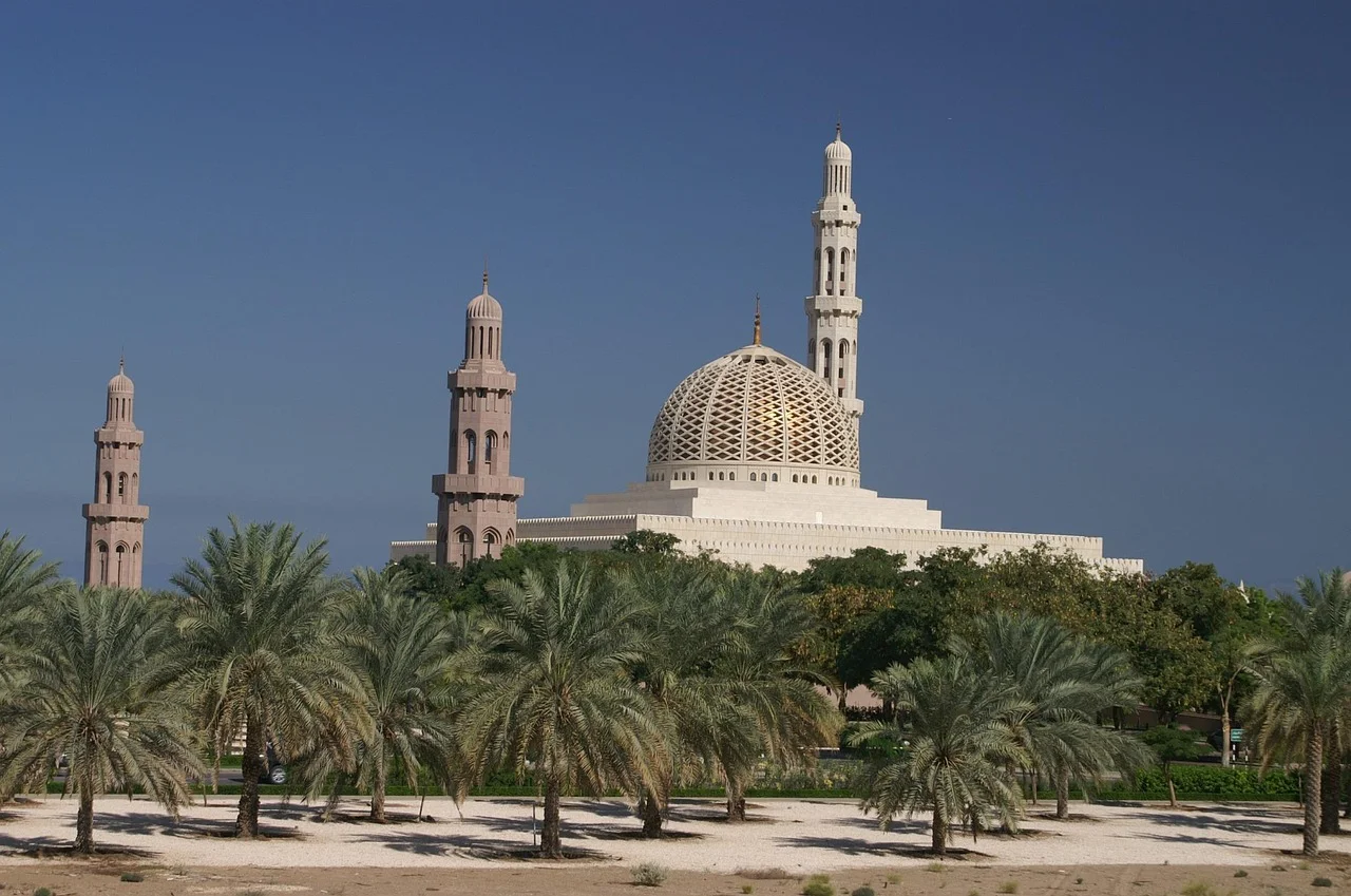 La Grande Mosquée Sultan Qaboos à Mascate, dôme doré et minarets blancs derrière une palmeraie de dattiers sous un ciel sans nuage