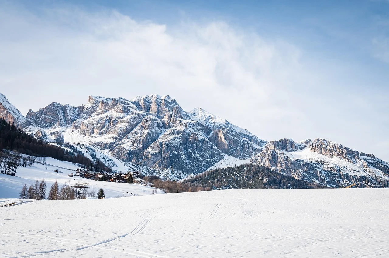 Massif rocheux des Dolomites couvert de neige au-dessus d'un plateau enneigé, hameau et mélèzes en contrebas