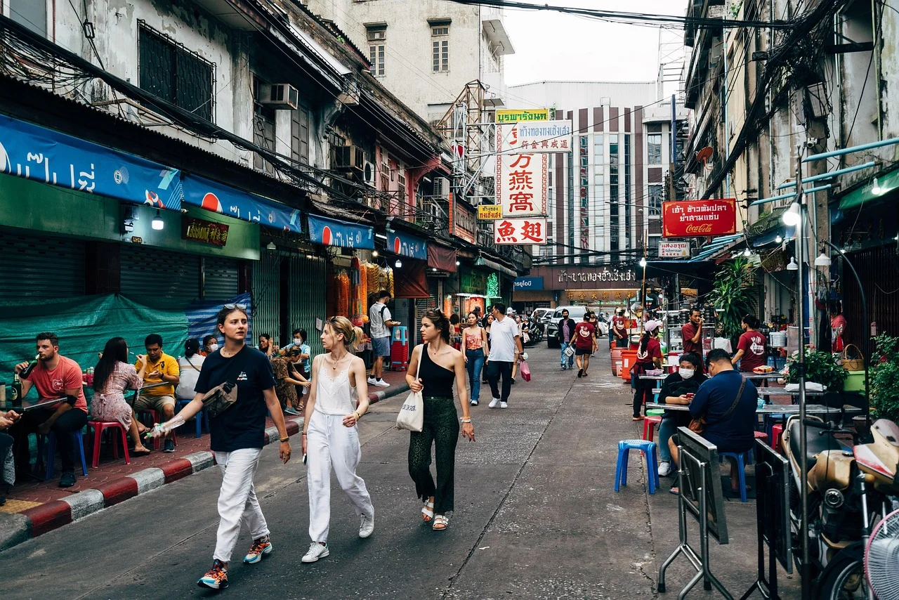 Passants dans une rue commerçante de Chinatown à Bangkok, bordée d'enseignes en thaï et en chinois et de tables de restaurants de rue.