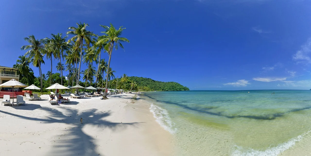Plage de sable blanc bordée de cocotiers et de parasols, eau turquoise et ciel bleu à Phu Quoc, au sud du Vietnam.