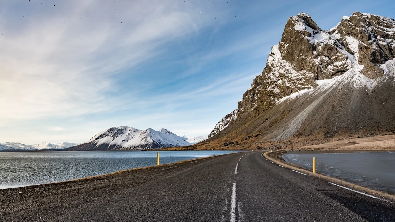 Route islandaise déserte longeant un fjord en hiver, avec des montagnes aux sommets enneigés sous un ciel bleu pâle.