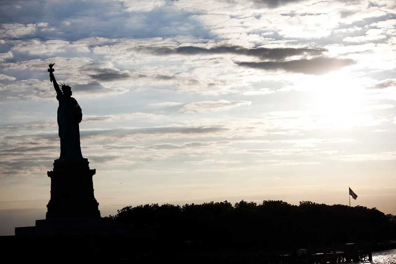 Silhouette de la statue de la Liberté à contre-jour sous un ciel nuageux, avec un drapeau américain au loin.