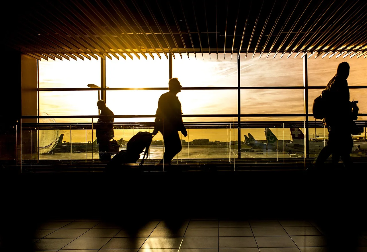 Silhouettes de voyageurs avec valises devant les baies vitrées d'un terminal d'aéroport au coucher du soleil.