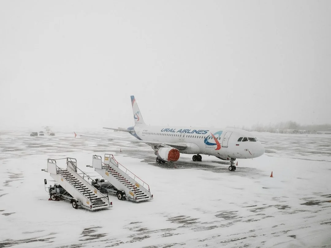 Avion de ligne stationné sur un tarmac enneigé sous un ciel blanc, deux passerelles mobiles couvertes de neige au premier plan.