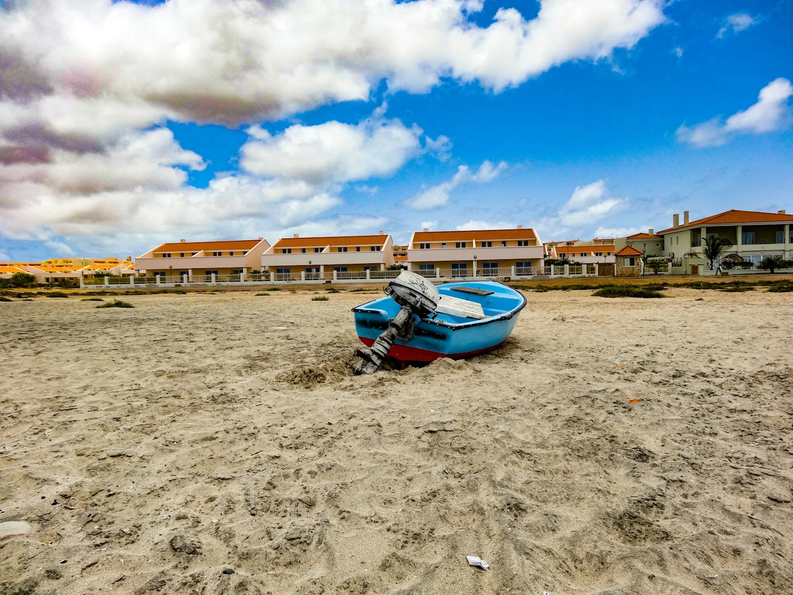 Barque de pêche bleue posée sur le sable de Sal au Cap-Vert, maisons basses sous un ciel bleu et nuages blancs