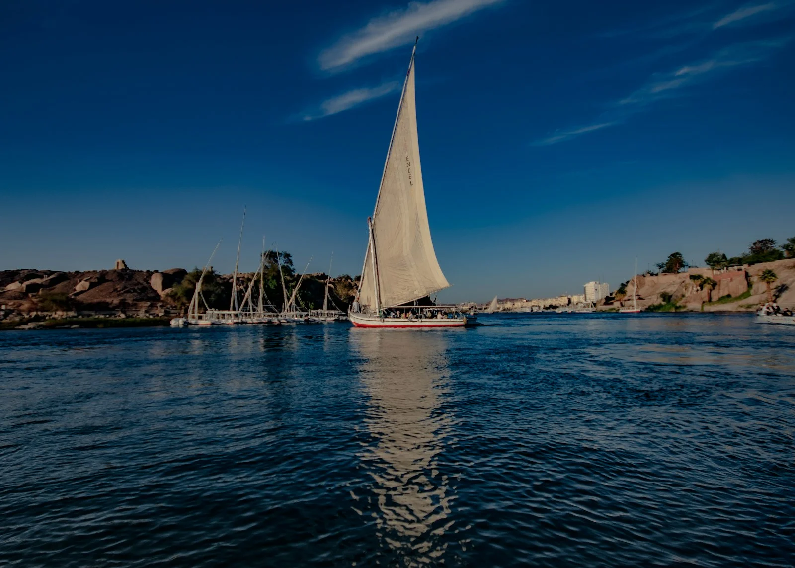 Felouque à voile blanche sur le Nil bleu, rochers de granit et palmiers sur la rive, à Assouan