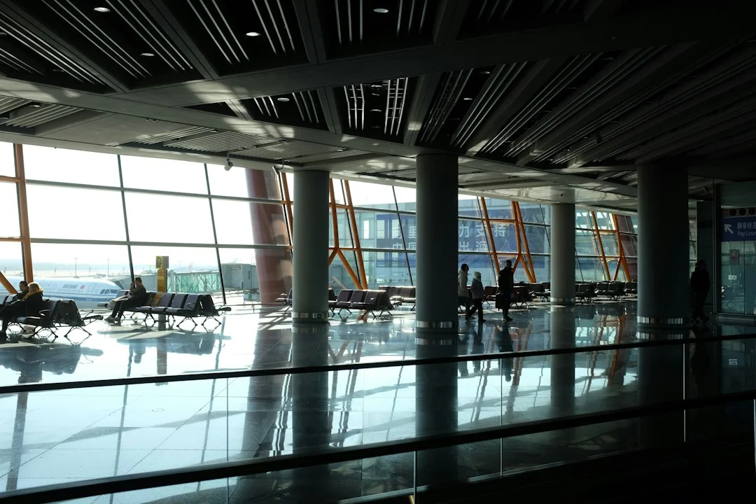 Salle d'embarquement d'aéroport presque vide, grandes baies vitrées sur le tarmac où stationne un avion, quelques passagers assis qui attendent.