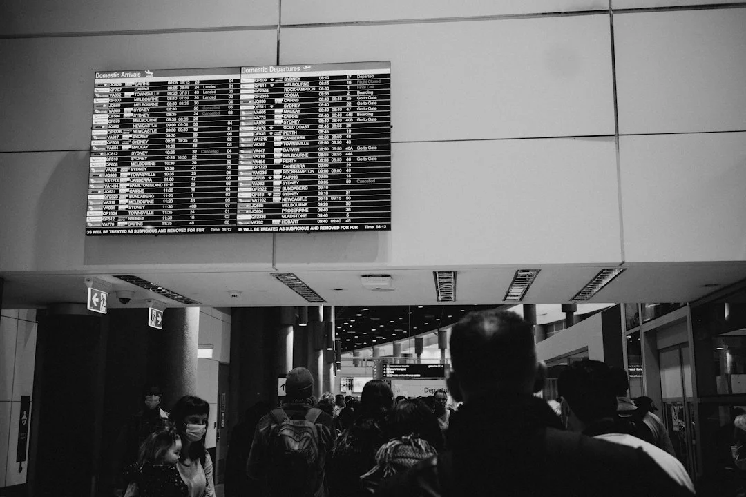 Tableau des départs et arrivées d'un aéroport, en noir et blanc, avec plusieurs vols marqués annulés au-dessus d'une foule de voyageurs.