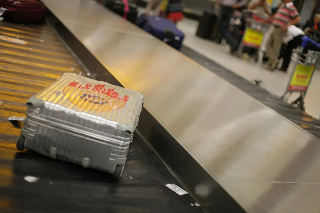 Valise rigide argentée posée seule sur un tapis à bagages d'aéroport, voyageurs et chariots flous à l'arrière-plan.