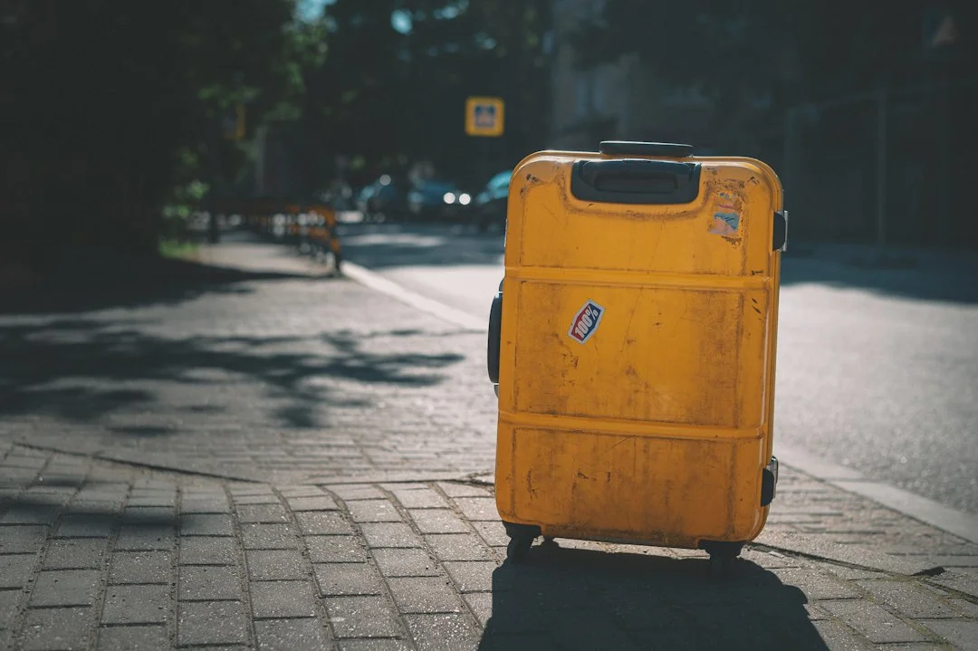 Valise rigide jaune très éraflée, avec des autocollants, posée sur un trottoir pavé au soleil.